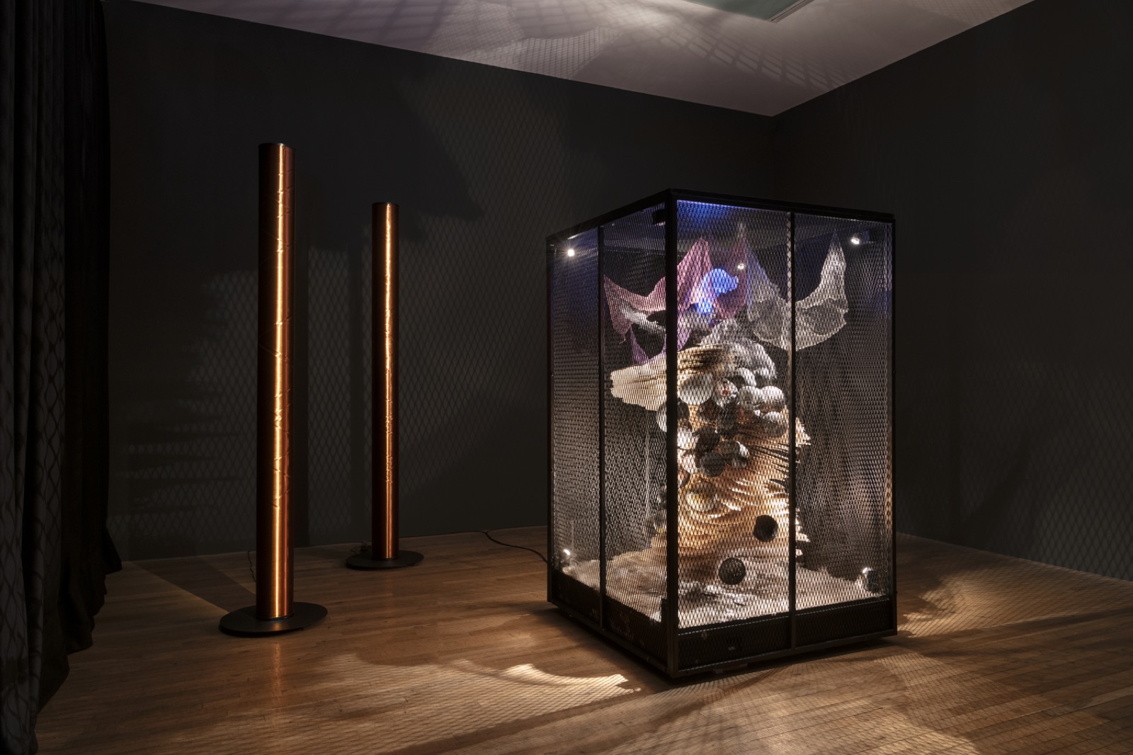 A dark gallery with two tall copper columns and a lit mesh cabinet containing a sculptural assemblage by Liliane Lijn.