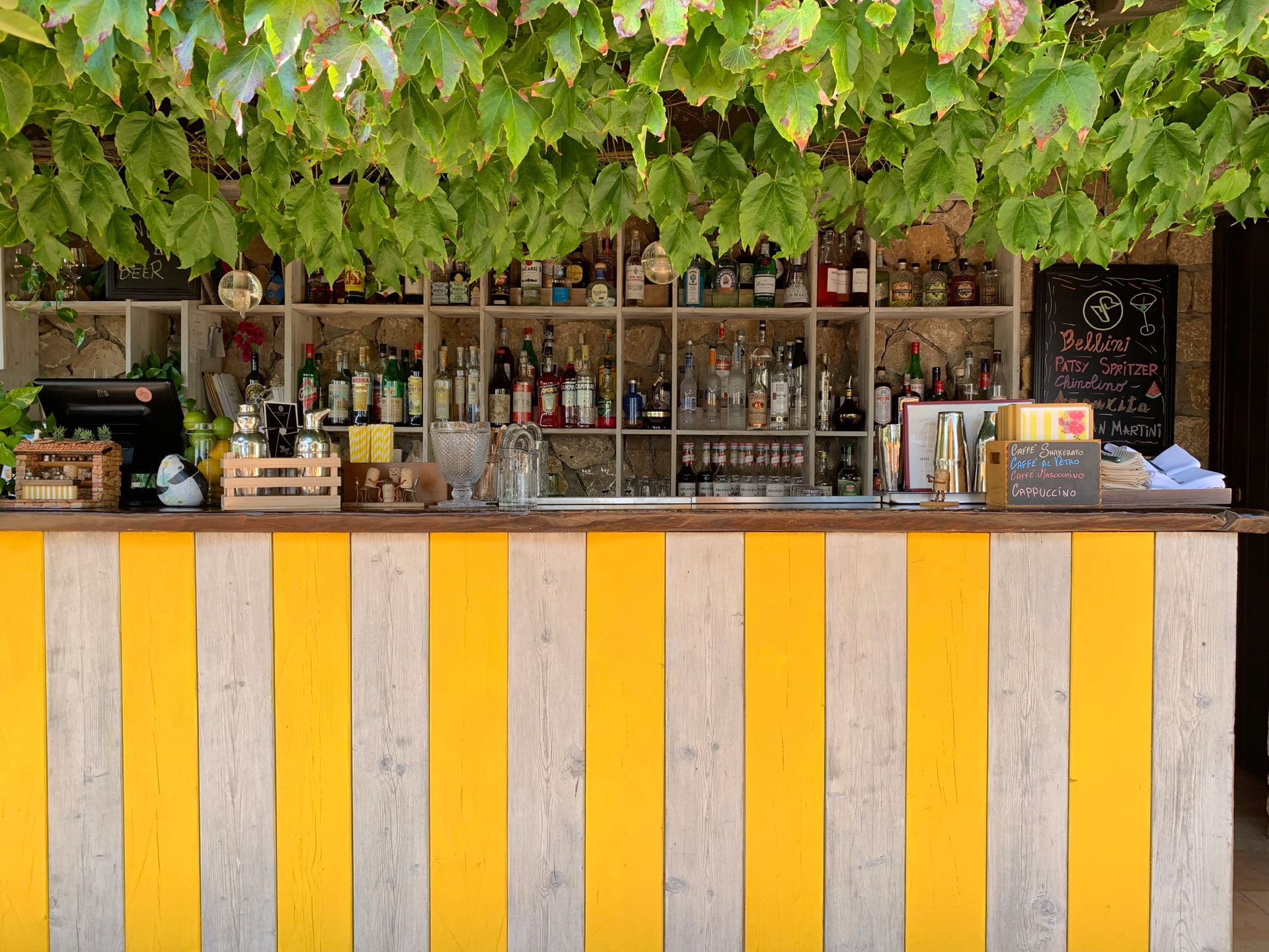 An outdoor bar with a yellow and white striped wooden counter under a vine canopy, shelves of bottles behind it and a chalkboard listing Bellini and Patsy Spritzer.