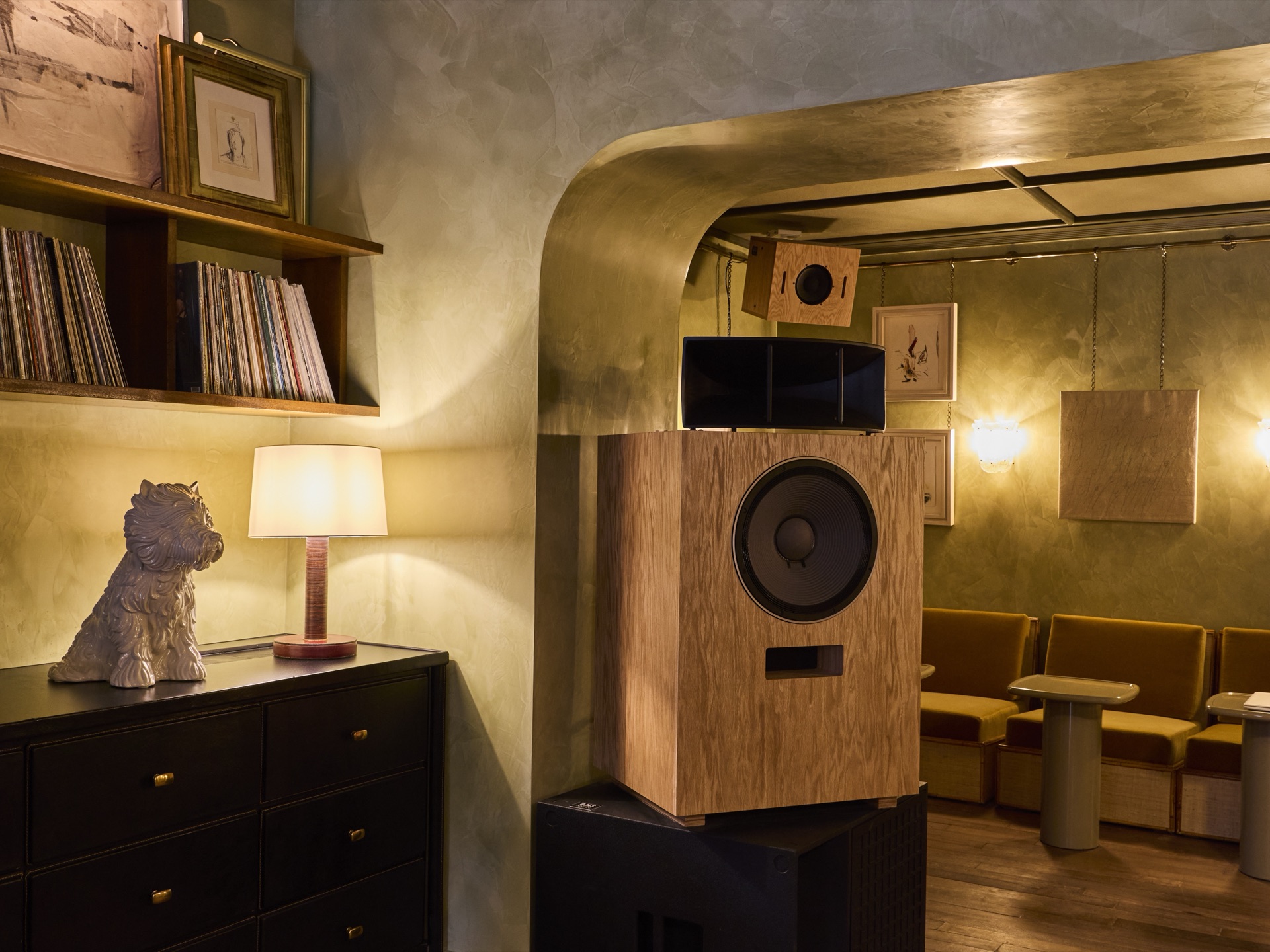 A large plywood loudspeaker on a black base in a room with green plaster walls, next to a shelf of vinyl records, framed drawings and a ceramic dog on a black chest of drawers.